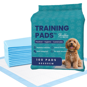 A package of Shabbee Dog Care training pads shown next to a stack of light blue absorbent pads on a white background. The packaging is teal with navy text and a photo of a small brown dog.